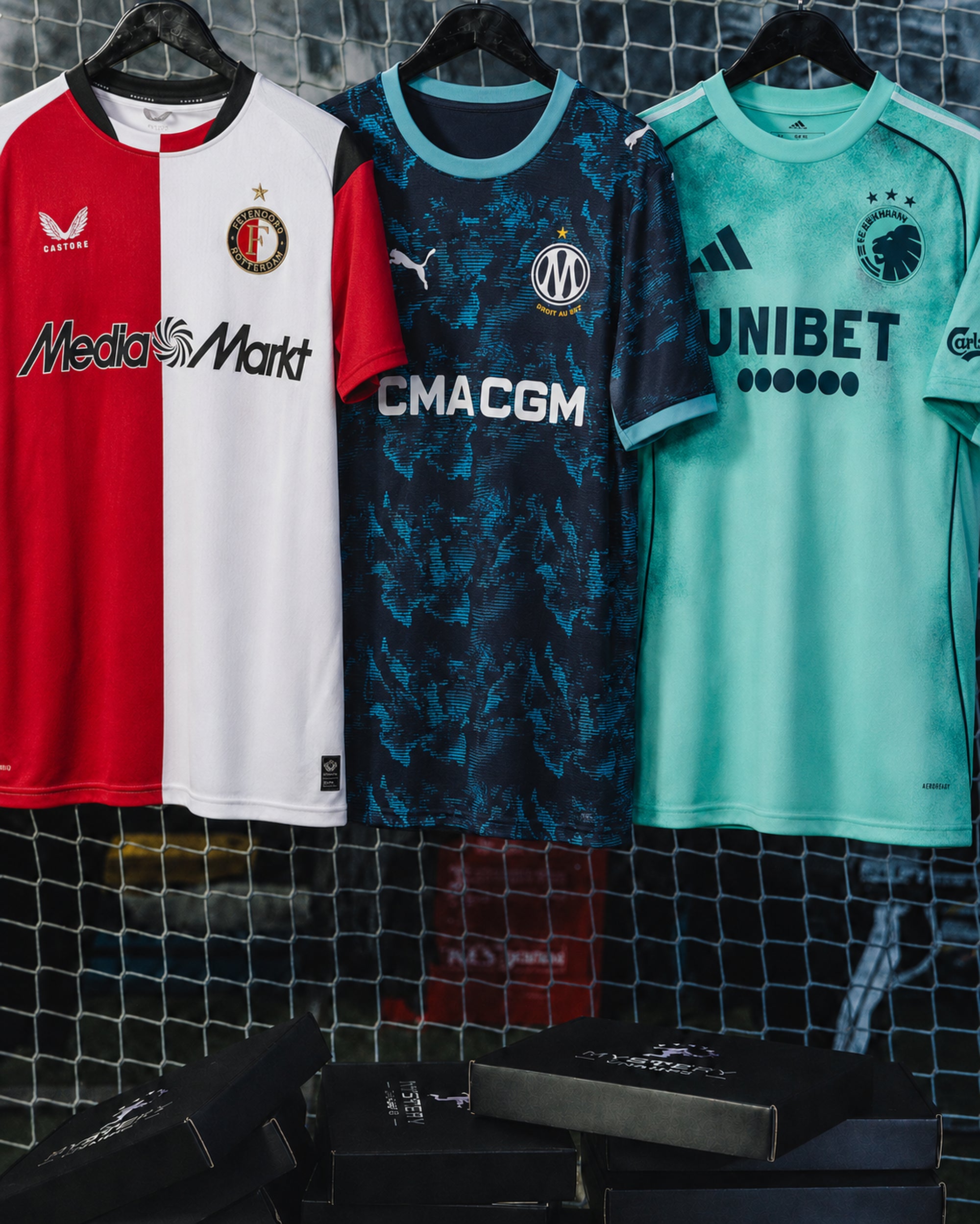 Drei Kinder-Fußballtrikots von Feyenoord, Olympique Marseille und FC Kopenhagen vor einem Tornetz mit Mystery-Champs-Boxen.